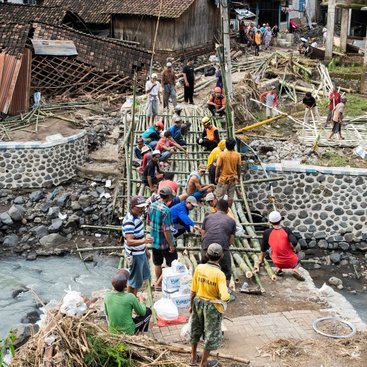 A group of people are working together to build a bridge across a river, some are standing on the bridge while others stand on either side of the riverbank